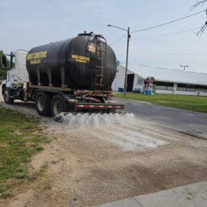 Seasonal Services A white truck with a large black tank labeled 'DUST CONTROL' is driving on a gravel road. The tank is dispensing a white substance onto the surface, with a building and green grass visible in the background.