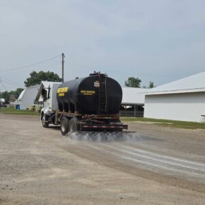 Seasonal Services A large black truck with a cylindrical tank marked "DUST CONTROL" is parked on a gravel road, releasing white dust control material onto the ground. In the background, a building with a white roof is visible alongside green grass.