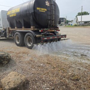 Seasonal Services A black tanker truck labeled 'DUST CONTROL' is parked on a gravel lot. Water is being dispersed from the rear of the truck, creating a sprinkling effect on the ground, while nearby structures and a dirt road are visible in the background.