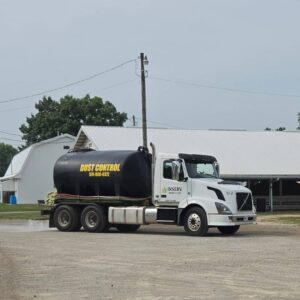 Seasonal Services A white and black truck with a large tank labeled "DUST CONTROL" parked on a gravel surface near agricultural buildings. The background features low-slung white structures, green trees, and power lines against a cloudy sky.