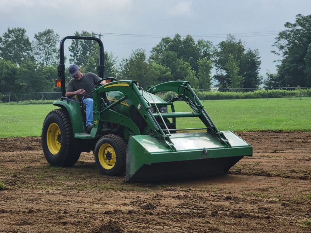 A man operates a green tractor with a front loader on a dirt field, with grassy land and trees in the background.
