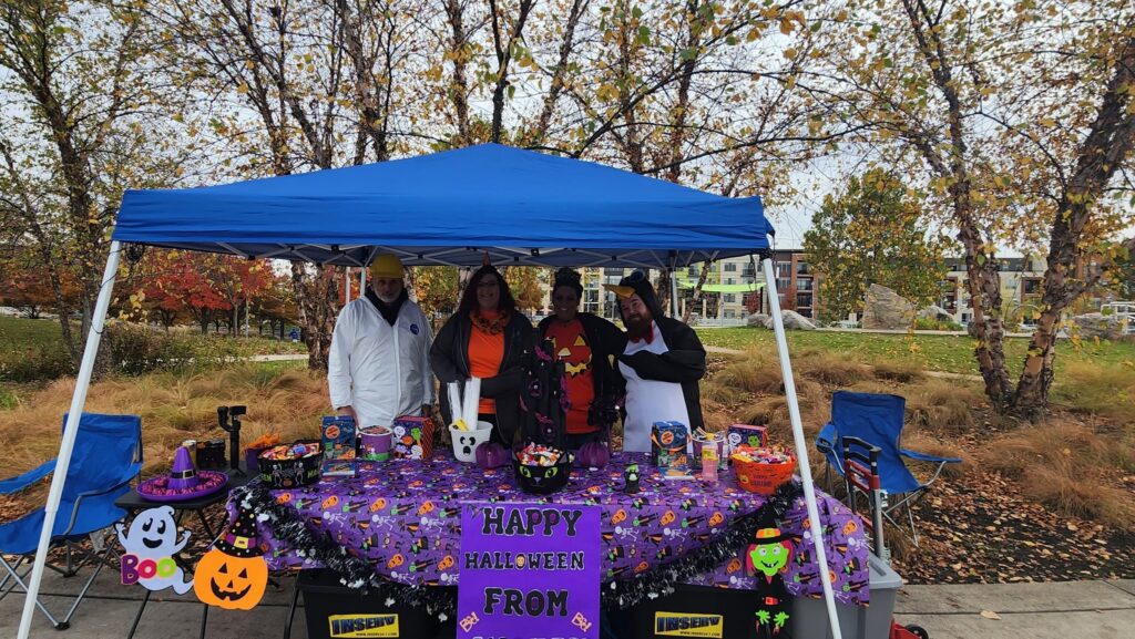 Four people stand behind a Halloween-themed table under a blue canopy tent in a park, with colorful decorations, candy, and a "Happy Halloween from INSERV" sign.
