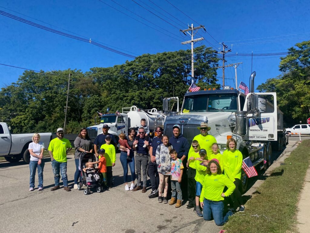 A group of people stands in front of a large tow truck on a roadside under a clear blue sky, with several American flags attached to the vehicle.