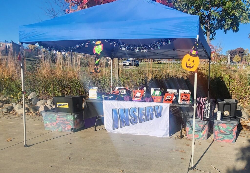 A blue pop-up canopy shelters a table with a white INSERV banner and colorful display items, with Halloween decorations and storage bins on a paved area.