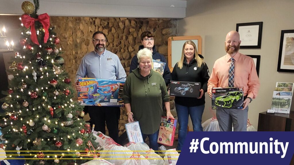 Five people stand indoors beside a decorated Christmas tree and wrapped gifts, with a large “#Community” banner in the lower right corner.