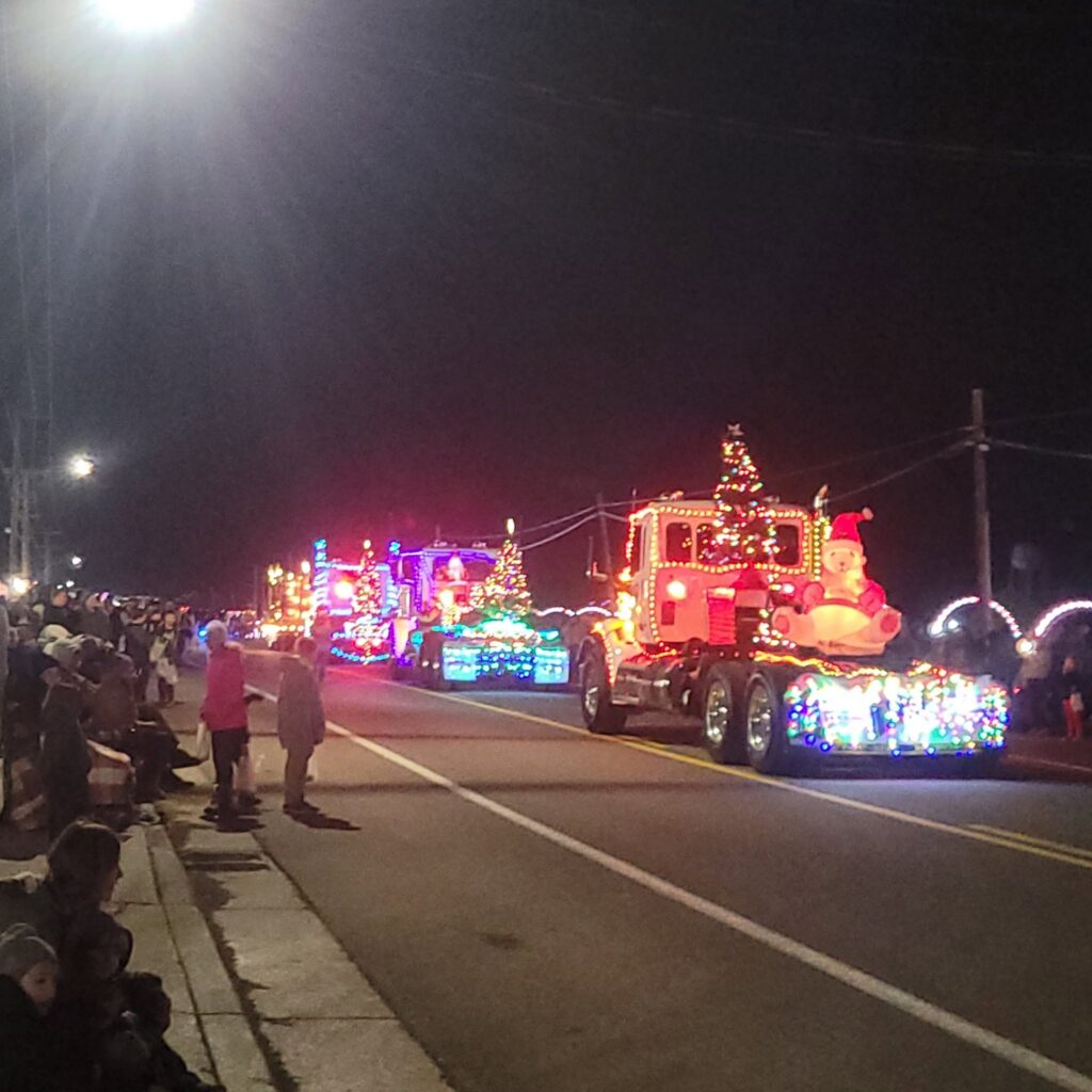 A Christmas parade float decorated with colorful lights, including a red truck with a Santa figure, moves down a nighttime street lined with spectators.