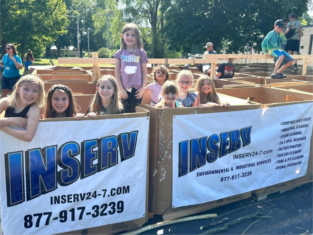 Several children stand inside a cardboard maze outdoors beside banners for INSERV, with trees and other people in the background.