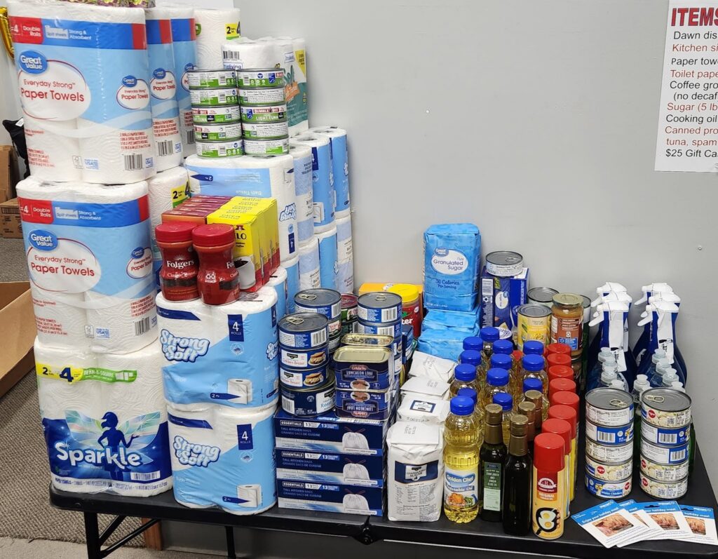 A table is covered with stacked household supplies and canned food, including paper towels, bottled oil, cleaning sprays, and boxed goods, with an item list posted on the wall behind it.