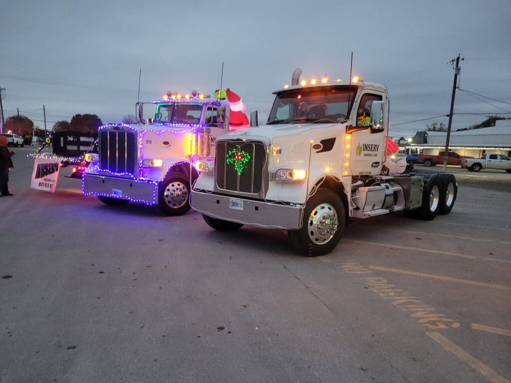 A white semi truck with orange marker lights and an Inserv logo on the door is parked beside a decorated truck outlined in colorful lights in a paved lot.