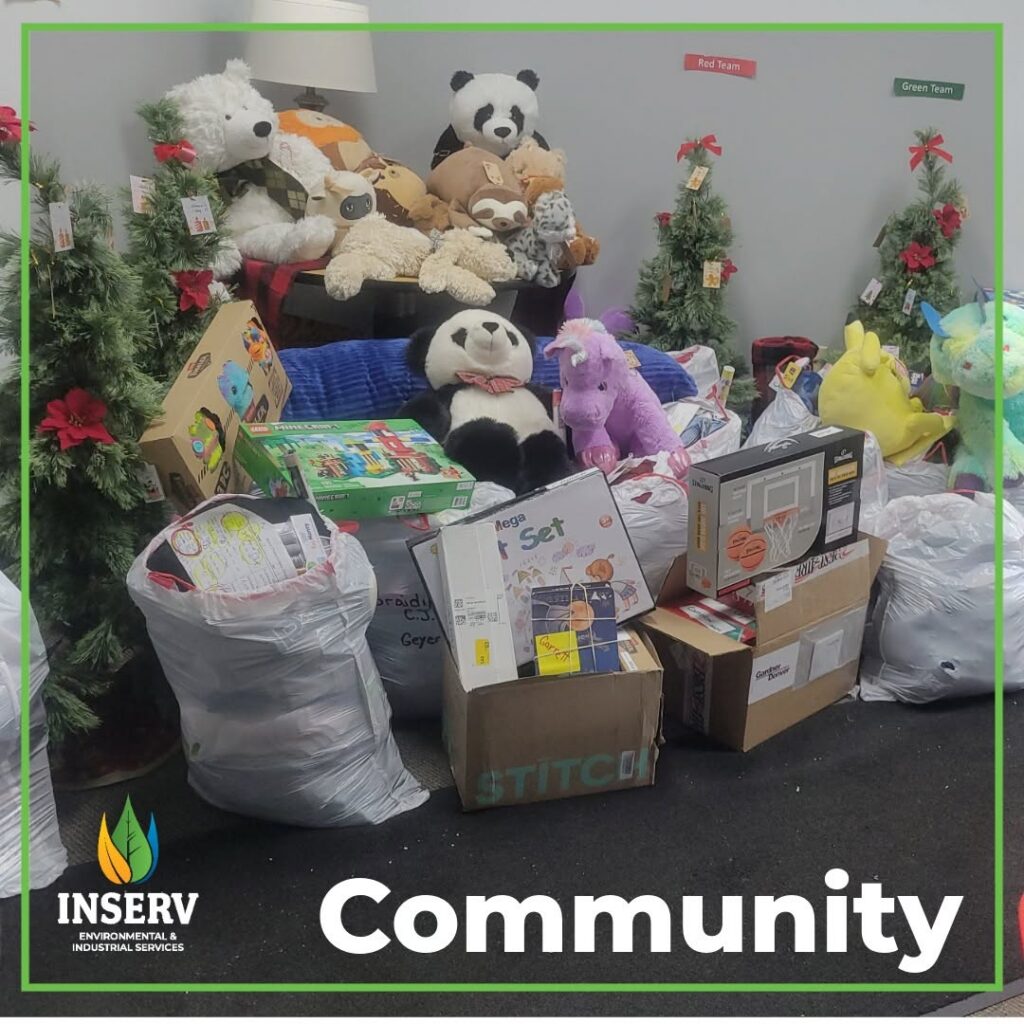 Large bags and cardboard boxes of donated toys and stuffed animals are arranged in front of small decorated Christmas trees with a "Community" sign.