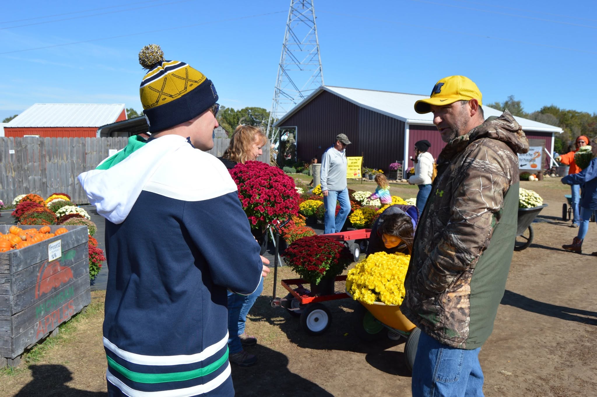 A man in a yellow cap and camouflage jacket stands beside a person in a blue-and-white jacket at an outdoor flower market with carts of potted chrysanthemums and a barn in the background.