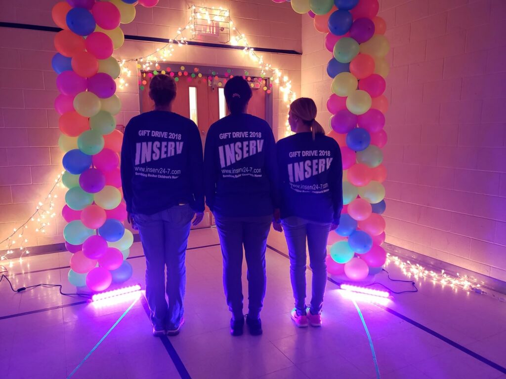 Three people in matching Gift Drive 2018 INSERV shirts stand beneath a colorful balloon arch and string lights outside double doors.
