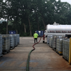 A worker in a high-visibility jacket stands beside long hoses between rows of large industrial liquid containers in an outdoor storage area with trees in the background.